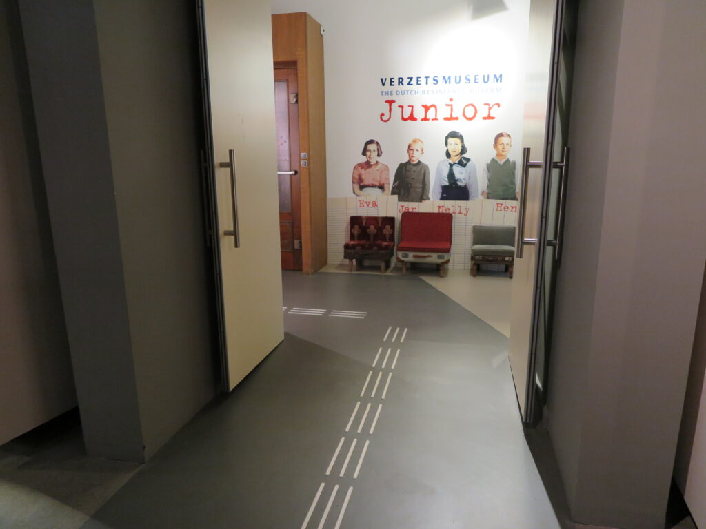 Op de foto staat de entree van Verzetsmuseum Junior, met een afbeelding van de hoofdpersonen Eva, Jan, Nelly en Henk op de muur.