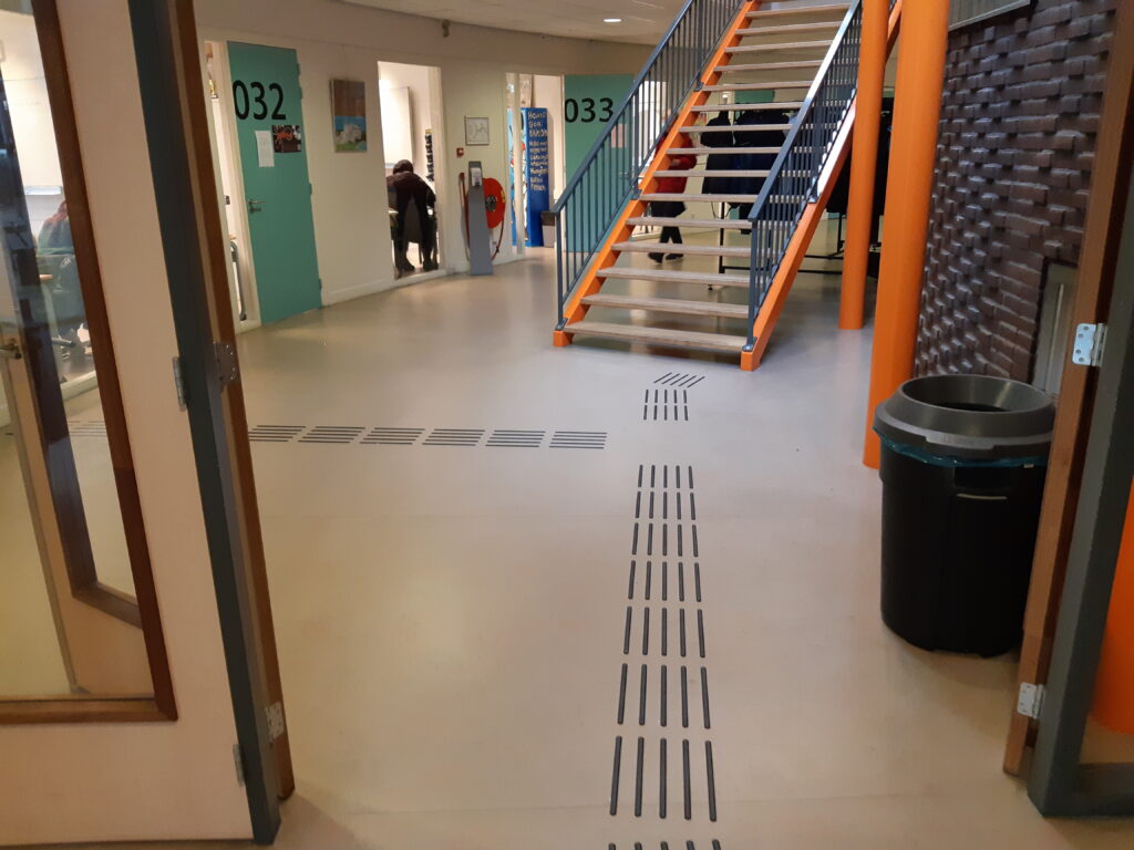 Geleideroute in een schoolgebouw. Zwarte ribbels op een lichte vloer. Op de achtergrond is een trap, links klaslokalen.