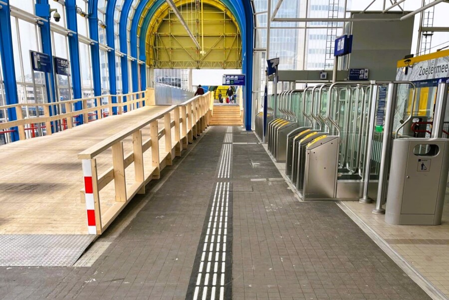 Route langs de checkin-checkout poorten. Links een houten talud en naar voren kijkend een grote gele boog aan het uiteinde van het overdekte gebied.