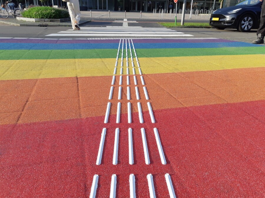 Zebrapad met regenboogkleuren voorzien van vijf witte blindegeleidelijnen naast elkaar. Laag vanaf de grond gefotografeerd.